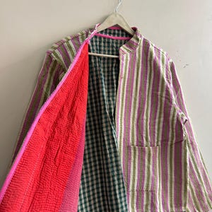 May include: A striped jacket in shades of green, pink, and white, with a red interior and a green and white plaid lining. The jacket has a mandarin collar and two pockets. The jacket is hanging on a wooden hanger.