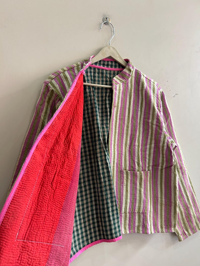 May include: A striped jacket in shades of green, pink, and white, with a red interior and a green and white plaid lining. The jacket has a mandarin collar and two pockets. The jacket is hanging on a wooden hanger.