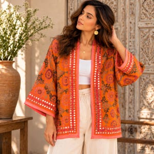 May include: An orange open-front kimono jacket featuring intricate floral embroidery in shades of pink, purple, and green, accented with small circular mirrors along the front placket and sleeve cuffs, worn over a white top and light-coloured trousers.