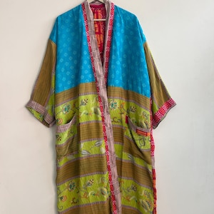May include: A long-sleeved kimono-style robe featuring a patchwork design with a bright blue upper section, a lime green lower section with floral patterns, and brown striped bands. The garment has a front opening with a patterned trim and two pockets.