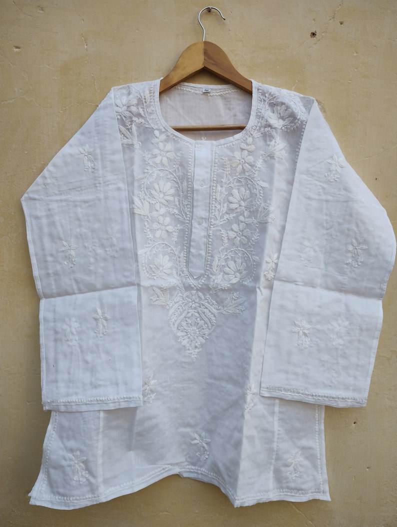 May include: White cotton tunic top with intricate floral embroidery. The top has a round neckline and long sleeves.