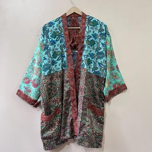 Pode incluir: Um quimono colorido com um padrão floral e paisley. A parte superior é turquesa com desenhos florais azuis e verdes, e a parte inferior apresenta um padrão paisley verde e vermelho. As mangas e as bordas são vermelhas.