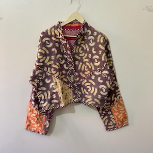 Puede incluir: Una chaqueta corta de color marrón, amarillo y naranja con cuello mandarín y mangas largas. La chaqueta está hecha de tela acolchada y tiene un diseño único de patchwork.