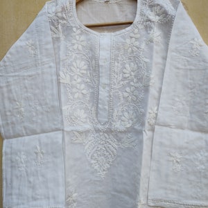 May include: White cotton tunic with intricate white embroidery featuring floral designs. The tunic has a round neckline and long sleeves.
