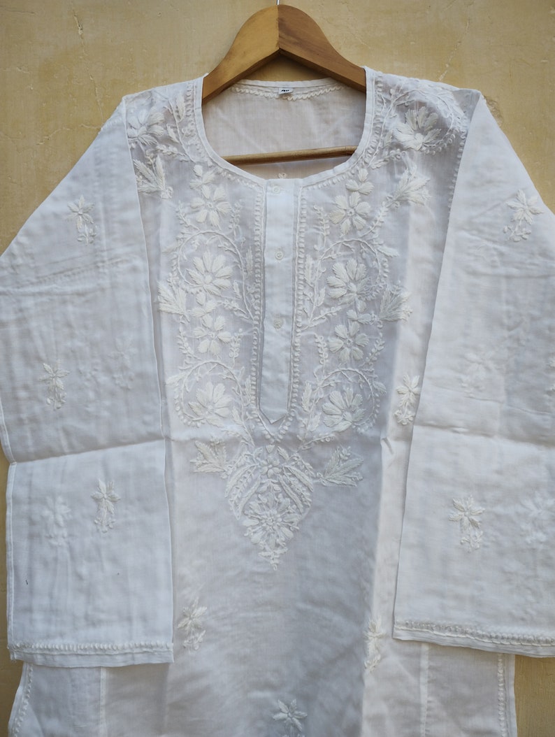 May include: White cotton tunic with intricate white embroidery featuring floral designs. The tunic has a round neckline and long sleeves.