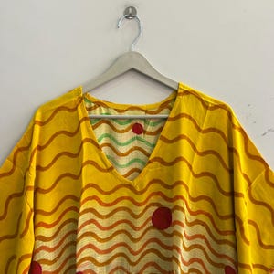 Pu&ograve; includere: Un caftano giallo con scollo a V e un motivo a righe ondulate arancioni e rosse. Il caftano presenta tre grandi cerchi rossi e un cordino in vita. &Egrave; appeso a un'appendiabiti grigio contro un muro bianco.