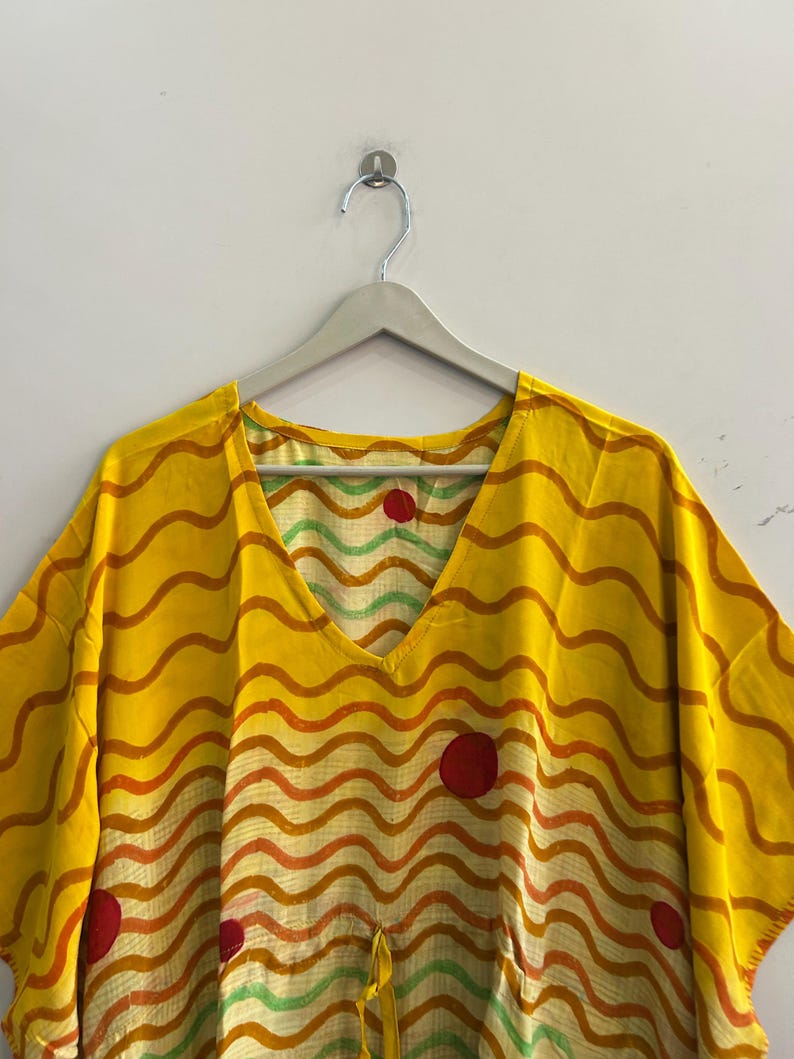 Pu&ograve; includere: Un caftano giallo con scollo a V e un motivo a righe ondulate arancioni e rosse. Il caftano presenta tre grandi cerchi rossi e un cordino in vita. &Egrave; appeso a un'appendiabiti grigio contro un muro bianco.