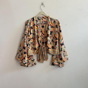 Pode incluir: Top com estampa paisley preta, laranja e amarela, com mangas compridas e largas. A parte superior é amarrada na cintura com uma fita de tecido combinando. Está pendurado em um cabide de madeira contra uma parede branca lisa.