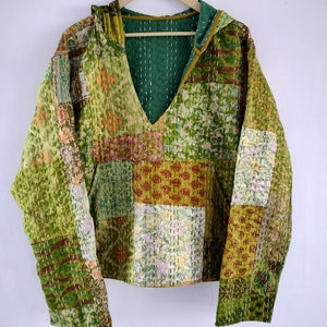 May include: A patchwork hoodie in shades of green, yellow, and pink, with a deep V-neck and a hood. The fabric is stitched with white thread, creating a quilted effect. The hoodie is hanging on a wooden hanger.