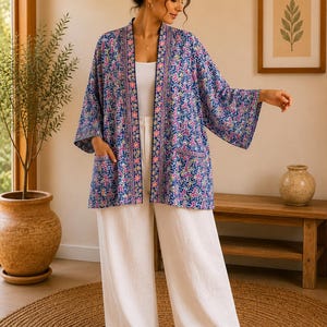 Può includere: Una giacca kimono blu aperta sul davanti, caratterizzata da un motivo geometrico ripetuto nei toni del rosa, bianco e blu scuro. Il capo presenta un bordo floreale a contrasto lungo l'apertura frontale e due tasche applicate.