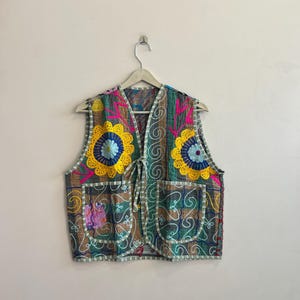 May include: A colorful, embroidered vest with a floral design. The vest features a mix of colors including yellow, blue, pink, and green. It has two front pockets and a tie closure. The vest is hanging on a hanger against a plain wall.