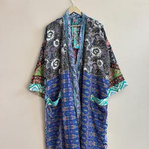 May include: A long, open-front robe with a patchwork design. The robe features a mix of floral and geometric patterns in shades of blue, purple, green, and pink. It has wide sleeves and two front pockets. The robe is hanging on a wooden hanger.