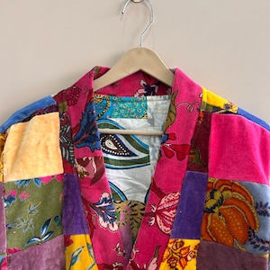May include: A patchwork robe with a vibrant mix of colors and patterns. The robe features a pink floral print on the collar and front, with various velvet squares in shades of pink, purple, green, and blue. It hangs on a wooden hanger.