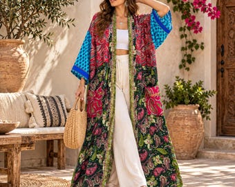 May include: A long, open-front kimono featuring a vibrant patchwork of paisley and floral patterns in shades of red, green, and black, with contrasting bright blue patterned sleeves, worn over a white crop top and wide-leg white trousers.