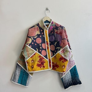 Può includere: Giacca patchwork colorata con collo alto, decorata con motivi floreali, a pois e a quadri. La giacca è appesa a un gancio di metallo su una parete bianca.