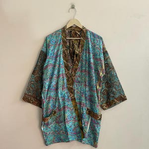 Puede incluir: Una bata kimono azul con estampado de cachemira y detalles en marrón y rosa. La bata tiene mangas anchas y dos bolsillos delanteros. Cuelga de una percha de madera sobre un fondo liso.