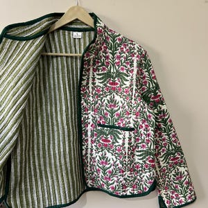 Może przedstawiać: Zielono-biała pikowana kurtka z kwiatowym nadrukiem po jednej stronie i paskowanym wzorem po drugiej. Kurtka ma zielony pasek i kieszeń po stronie z kwiatami.