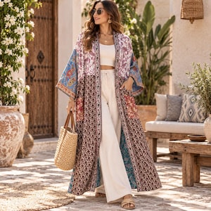May include: A long, open-front kimono featuring a patchwork design with various floral and paisley patterns in shades of blue, brown, white, and red. The garment has wide sleeves and two front pockets, worn over a white crop top and white trousers.