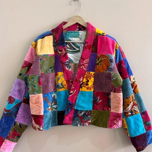 May include: A patchwork jacket with a vibrant mix of colors and patterns. The jacket features a variety of fabrics in squares of pink, blue, green, yellow, and purple. It is hanging on a wooden hanger.