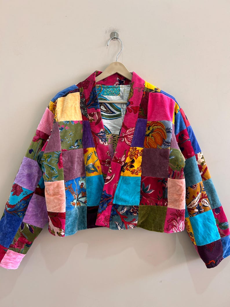May include: A patchwork jacket with a vibrant mix of colors and patterns. The jacket features a variety of fabrics in squares of pink, blue, green, yellow, and purple. It is hanging on a wooden hanger.