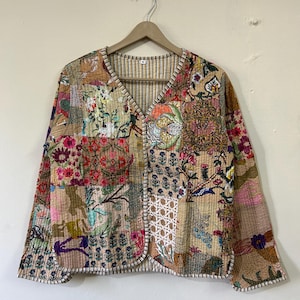 Op de afbeelding: Een beige gewatteerd jack met een patchwork-ontwerp met bloemen- en geometrische patronen in verschillende kleuren, waaronder roze, rood, groen en blauw. Het jack heeft een V-hals en lange mouwen.