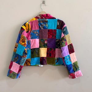 May include: A patchwork jacket with a vibrant mix of colors and patterns. The jacket features a variety of fabrics in squares of pink, blue, green, purple, and yellow. The jacket is hanging on a white hanger.