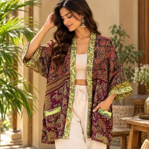 Op de afbeelding: Een kimono-jas met lange mouwen en een open voorkant, voorzien van een donkerpaarse basis met een veelkleurig paisley-patroon. Het kledingstuk heeft een contrasterende lichtgroene rand met patroon langs de opening, mouwen en twee zakken.