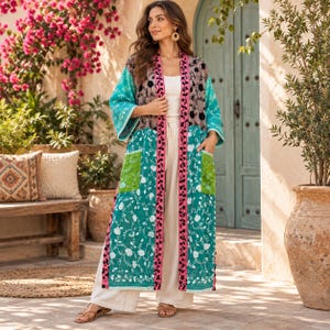 May include: A long, open-front kimono-style duster featuring a mix of teal, green, and grey panels with white floral embroidery. The garment includes a prominent black vertical trim with floral detailing, two large patch pockets, and long sleeves.