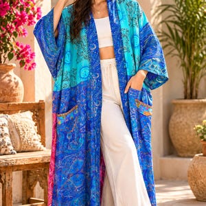 May include: A long, open-front kimono robe in vibrant shades of blue, featuring a celestial print with sun and moon motifs, intricate paisley patterns, wide sleeves, and two front patch pockets, worn over a white crop top and white wide-leg trousers.