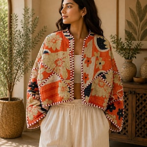 Puede incluir: Una chaqueta acolchada corta y abierta por delante con un diseño de patchwork en tonos naranja, crema y azul oscuro con motivos florales. La prenda tiene mangas largas y anchas, con un borde a rayas rojas y blancas en los bordes y el cuello.