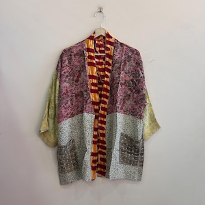 May include: A patchwork kimono-style jacket with a vibrant mix of colors and patterns. The jacket features a burgundy and pink floral print, a gray and white speckled pattern, and yellow sleeves. It has a striped orange and burgundy front panel and two pockets.