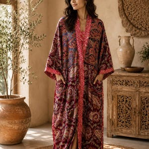 Könnte beinhalten: Ein bodenlanger Kimono-Bademantel mit einer tiefburgunderroten Basis und einem mehrfarbigen Paisley-Muster in Blau-, Orange- und Goldtönen. Das Kleidungsstück verfügt über lange, weite Ärmel, zwei Vordertaschen und eine kontrastierende rote Bordüre.