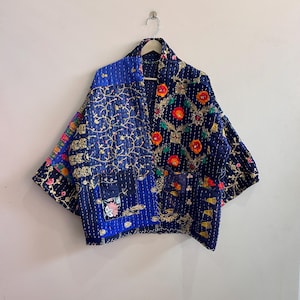 Peut inclure: Veste patchwork bleue ornée de broderies florales rouges, oranges et dorées. La veste est suspendue à un cintre en bois. Elle présente une texture matelassée et un design unique, contre un mur blanc.