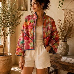 May include: A cropped, long-sleeve jacket featuring a vibrant, multi-coloured floral watercolour print in shades of pink, orange, and green, finished with dark blue piping along the edges and seams, worn over a cream-coloured tank top and shorts.