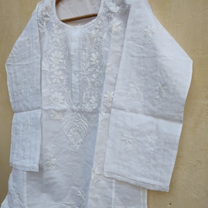 May include: White cotton tunic top with delicate white embroidery. The top has a round neckline and long sleeves.