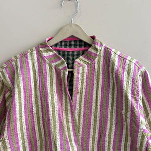 May include: A striped jacket in shades of pink, green, and white, hanging on a wooden hanger. The jacket features a mandarin collar and a quilted design. The interior lining is a plaid pattern with pink trim.