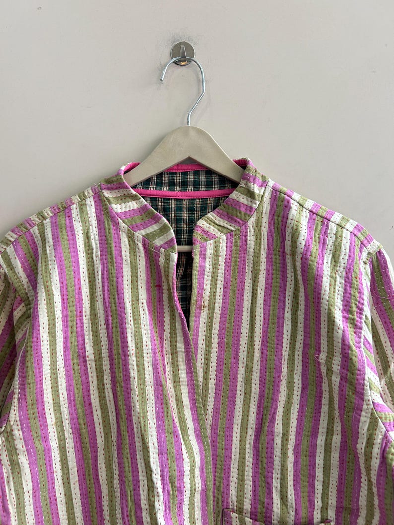 May include: A striped jacket in shades of pink, green, and white, hanging on a wooden hanger. The jacket features a mandarin collar and a quilted design. The interior lining is a plaid pattern with pink trim.
