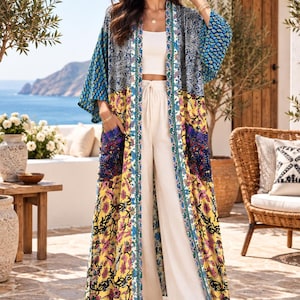 Può includere: Un kimono lungo aperto sul davanti con un design patchwork caratterizzato da motivi floreali e paisley nei toni del blu, giallo e viola. Il capo presenta maniche ampie, due tasche frontali e un bordo verticale lungo l'apertura, indossato sopra un top e pantaloni bianchi.