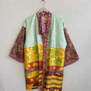 May include: A patchwork kimono-style jacket featuring a mix of fabrics with vibrant yellow, teal, and deep maroon patterns. The garment includes long sleeves, a front tie closure, and a colorful floral and geometric print design.