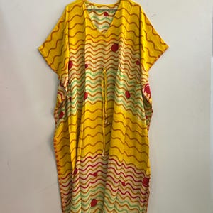 Pu&ograve; includere: Un caftano giallo con scollo a V e maniche corte, appeso a una gruccia di legno. Il caftano presenta un motivo ondulato a righe rosse, arancioni e verdi, con pois rossi. Un cordoncino stringe la vita. Il capo &egrave; realizzato in tessuto leggero.