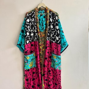 May include: A long-sleeved kimono-style robe featuring a patchwork design with vibrant panels of black, bright pink, and turquoise fabric. The garment is adorned with intricate floral embroidery throughout and includes two front pockets and a patterned trim.