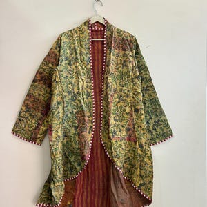 Peut inclure: Une longue veste ouverte sur le devant avec un motif floral dans les tons de jaune, de vert et de marron. La veste a une bordure rayée rouge et blanche et une doublure rayée rouge et marron. La veste est suspendue à un mur blanc.