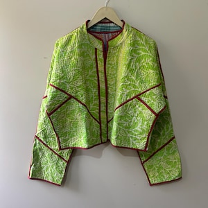 May include: A lime green quilted jacket with a white pattern and red trim. The jacket is hanging on a wooden hanger. The jacket has a round collar and long sleeves.