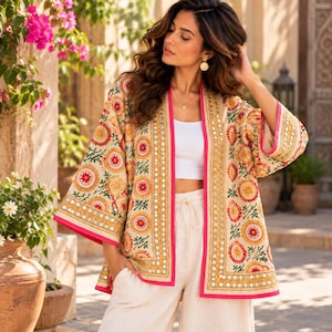 Könnte beinhalten: Eine beige, vorne offene Kimono-Jacke mit lebendigen kreisförmigen Stickereien in Rot, Orange und Gelb, akzentuiert durch kleine reflektierende Spiegelverzierungen an den Vorderteilen und Ärmeln, eingefasst mit leuchtend pinkfarbenen Kanten.