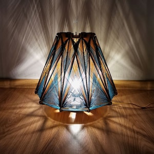 Pode incluir: Uma luminária de mesa geométrica com estrutura de madeira e cúpula feita de fios azuis, dourados e pretos. A luminária está acesa, projetando luz e sombras no chão e na parede de madeira.