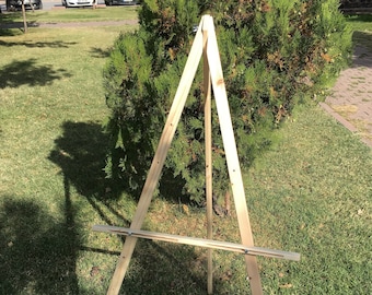 May include: A wooden easel with a natural finish. The easel is made of three pieces of wood that are joined together to form a triangular shape. The easel is standing on a grassy surface.