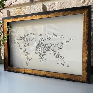 May include: A black frame with a gold trim surrounds a white map of the world. The map shows the continents and countries in black lines.
