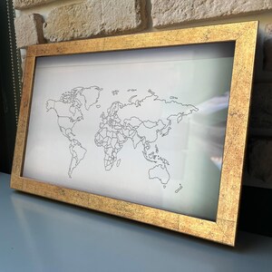 May include: A rectangular wall art piece featuring a black line drawing of a world map on a white background, encased in a textured, metallic gold frame with a distressed finish. The map displays continental outlines and country borders.