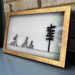 May include: A framed wall art piece featuring a black line drawing of three people walking with luggage toward a signpost. The signpost displays the text: LONDRES, NEW YORK, PARIS, BANGKOK, and RIO. The frame has a textured, metallic gold finish.