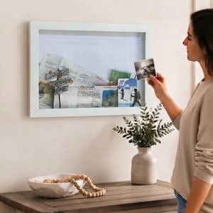 May include: A white shadow box frame mounted on a wall, containing various travel mementos and photographs. The frame features a deep interior space for displaying items behind a clear front panel, providing a structured way to preserve memories.
